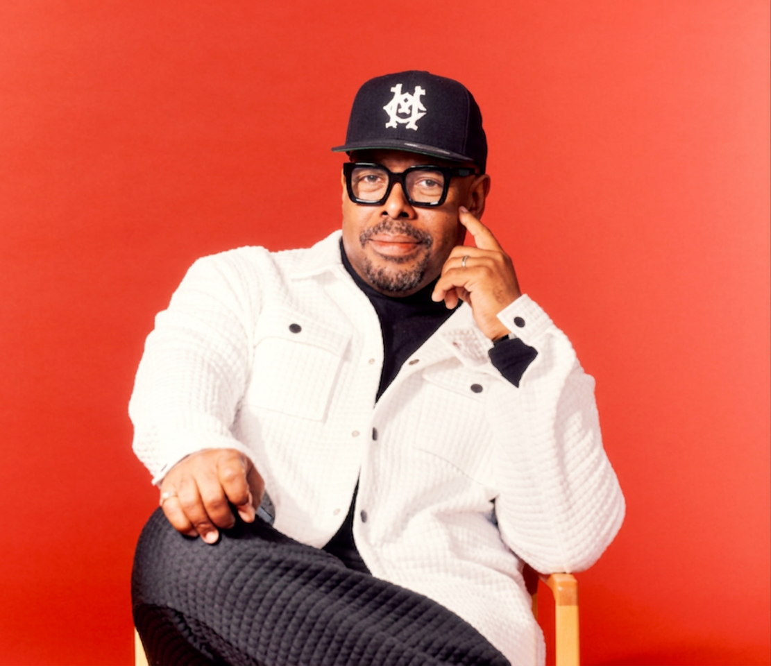 A man wearing a black cap, glasses, and a white button-up shirt, seated against a bright red background, resting his chin on his hand.