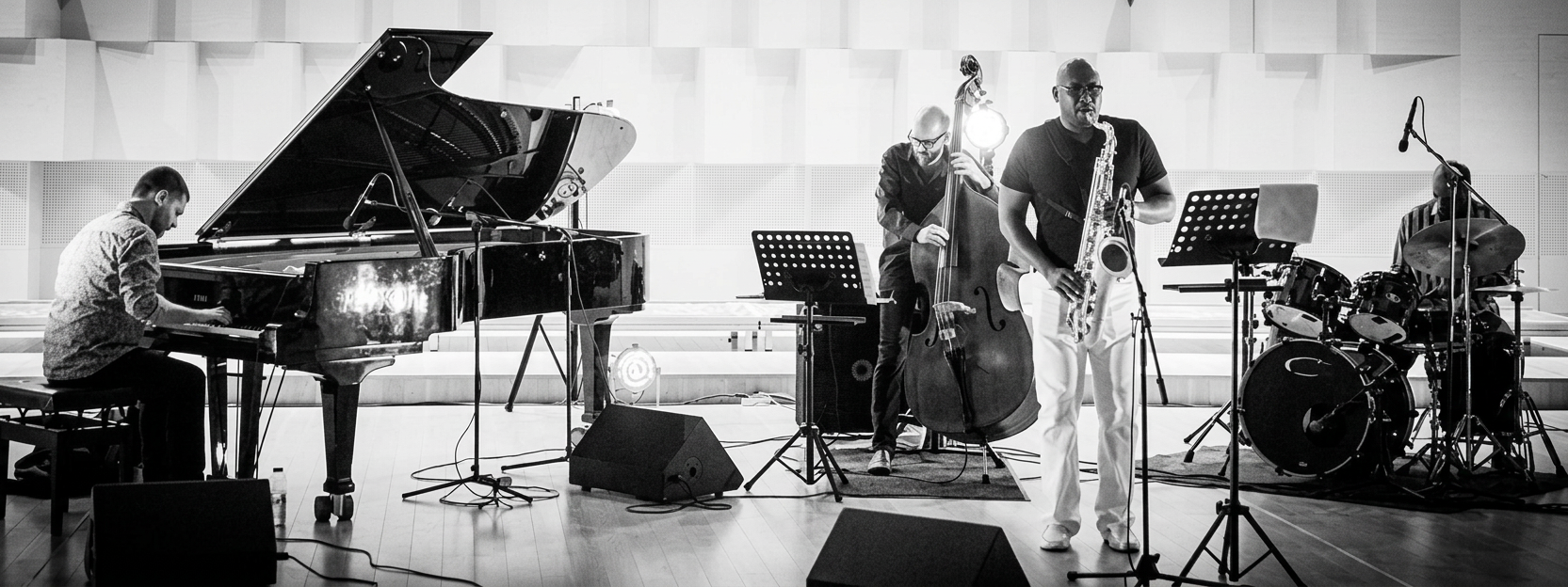 Elevation A jazz band performing on stage, featuring a pianist, saxophonist, bassist, and drummer, with musical instruments and sheet music visible.